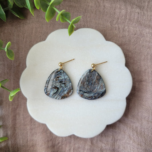 Top view of a pair of gray, brown guitar pick shaped earrings displayed on a white round scalloped tray.