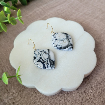 Left side view of a pair of black, white marble round diamond shaped earrings displayed on a white round scalloped tray.