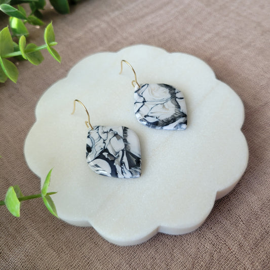 Left side view of a pair of black, white marble round diamond shaped earrings displayed on a white round scalloped tray.