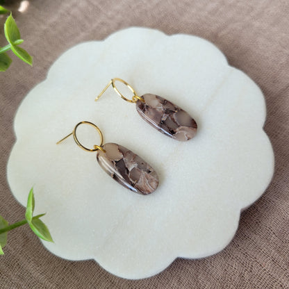 Left side view of a pair of brown marble long oval earrings displayed on a white round scalloped tray.