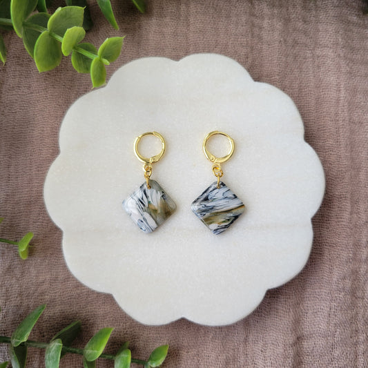 Top view of a pair of black, white, brown, moss square shaped earrings displayed on a white round scalloped tray.