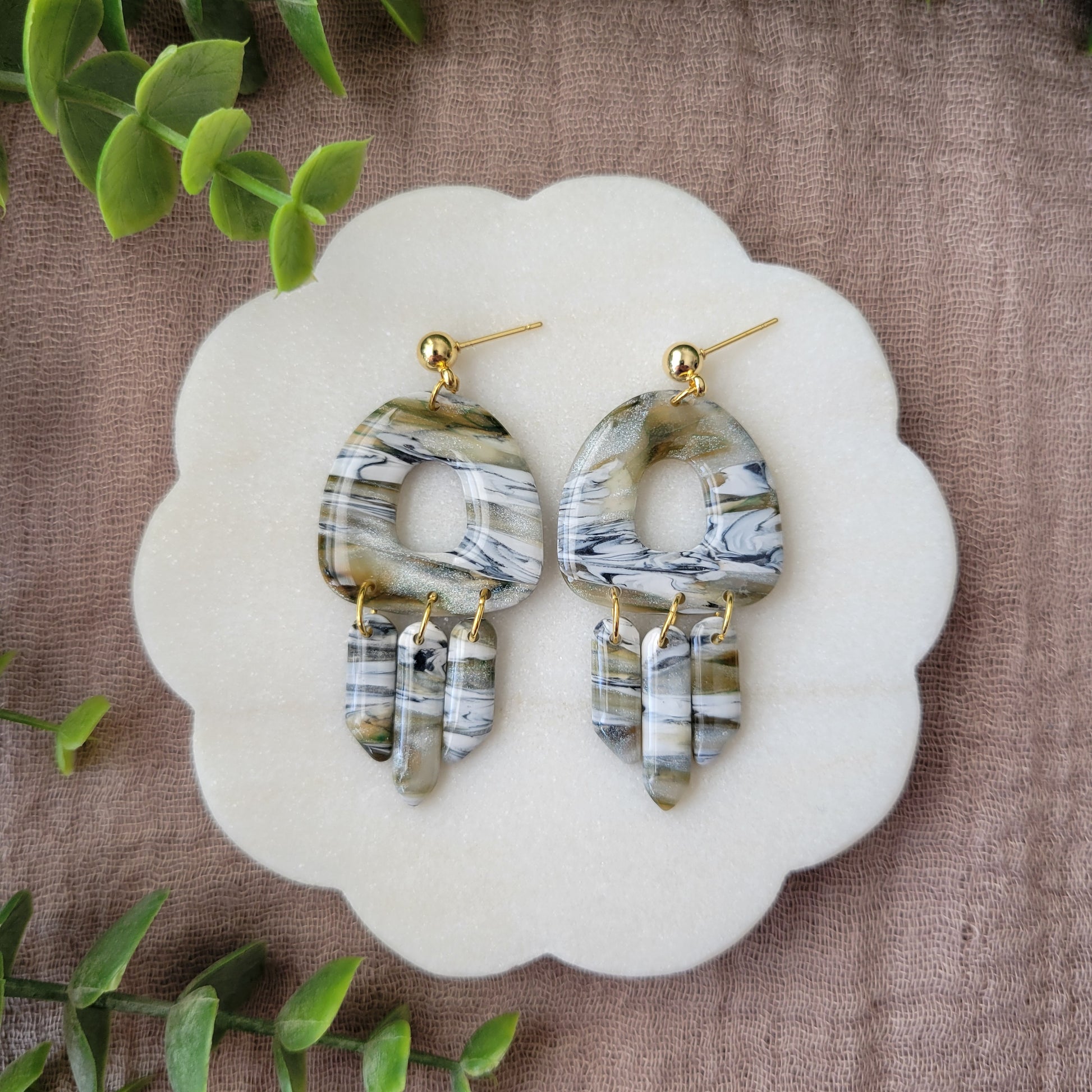Top view of a pair of green, white, black, brown marbled earrings displayed on a white round scalloped tray.
