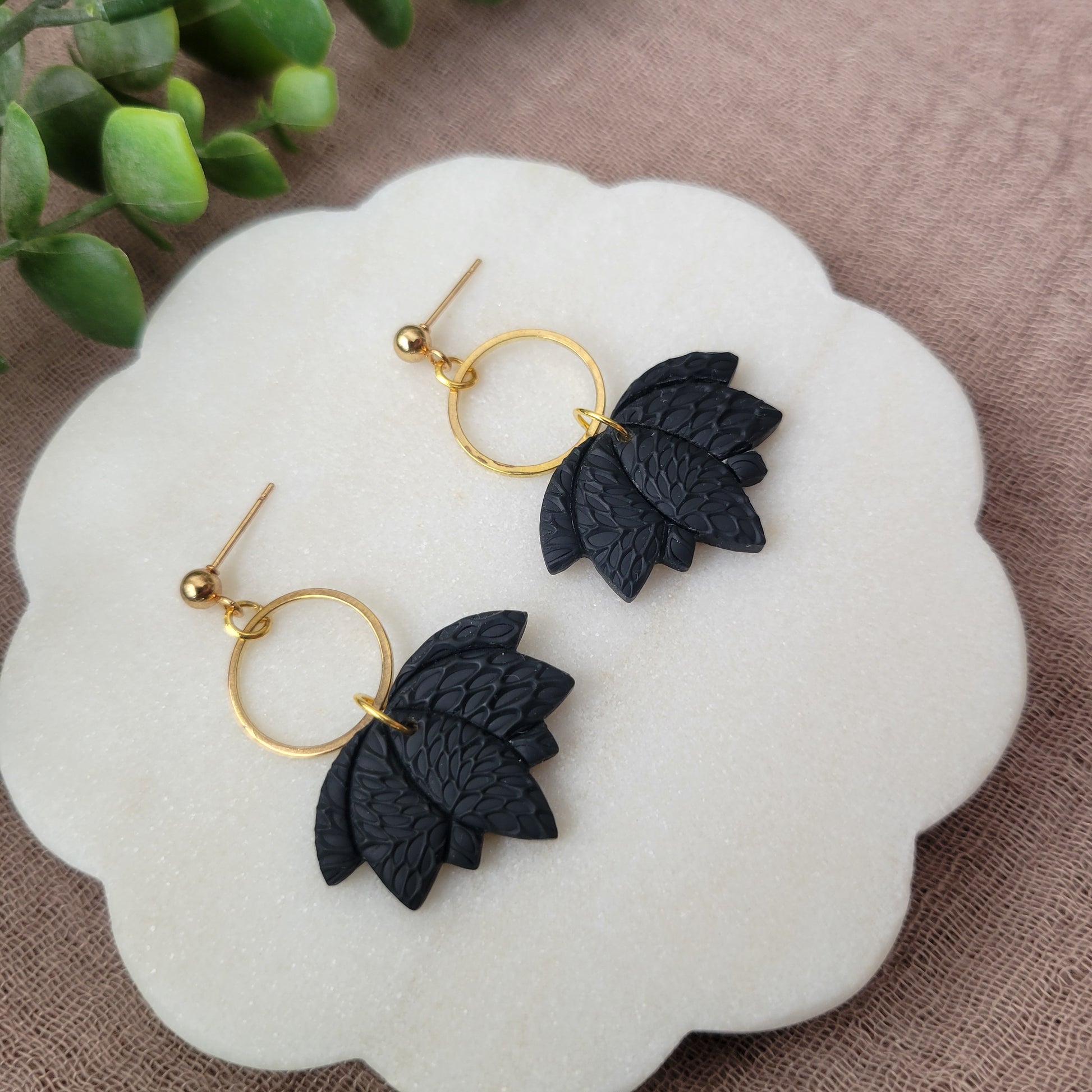Left side view of a pair of black lotus earrings with interwoven leaves texture and gold circle bezel displayed on a white round scalloped tray.