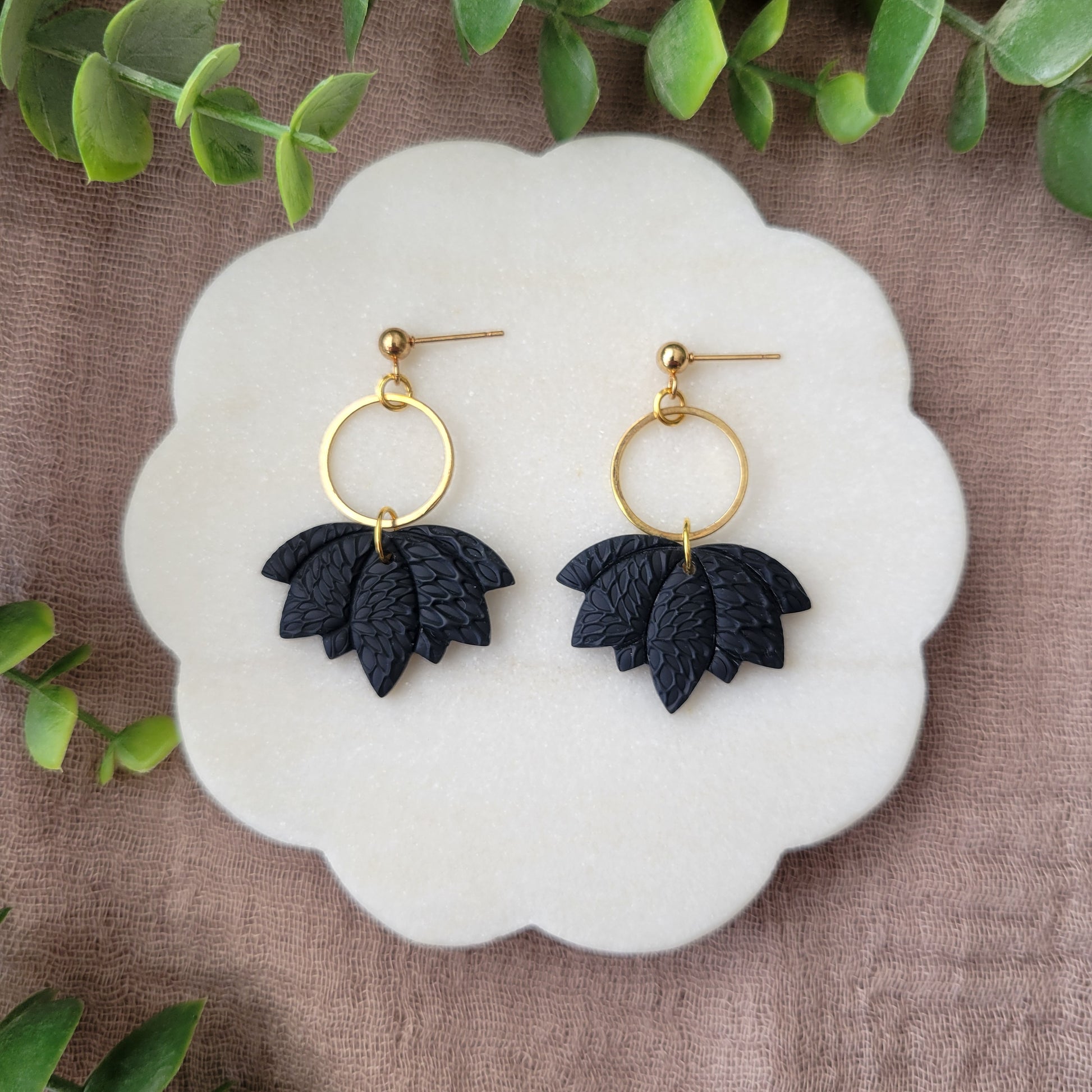 Top view of a pair of black lotus earrings with interwoven leaves texture and gold circle bezel displayed on a white round scalloped tray.