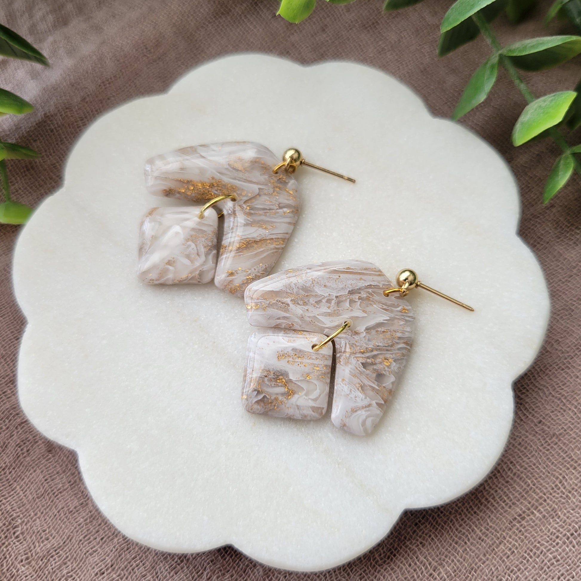Right side view of a pair of white, gold marble boomerang shaped earrings displayed on a white round scalloped tray.