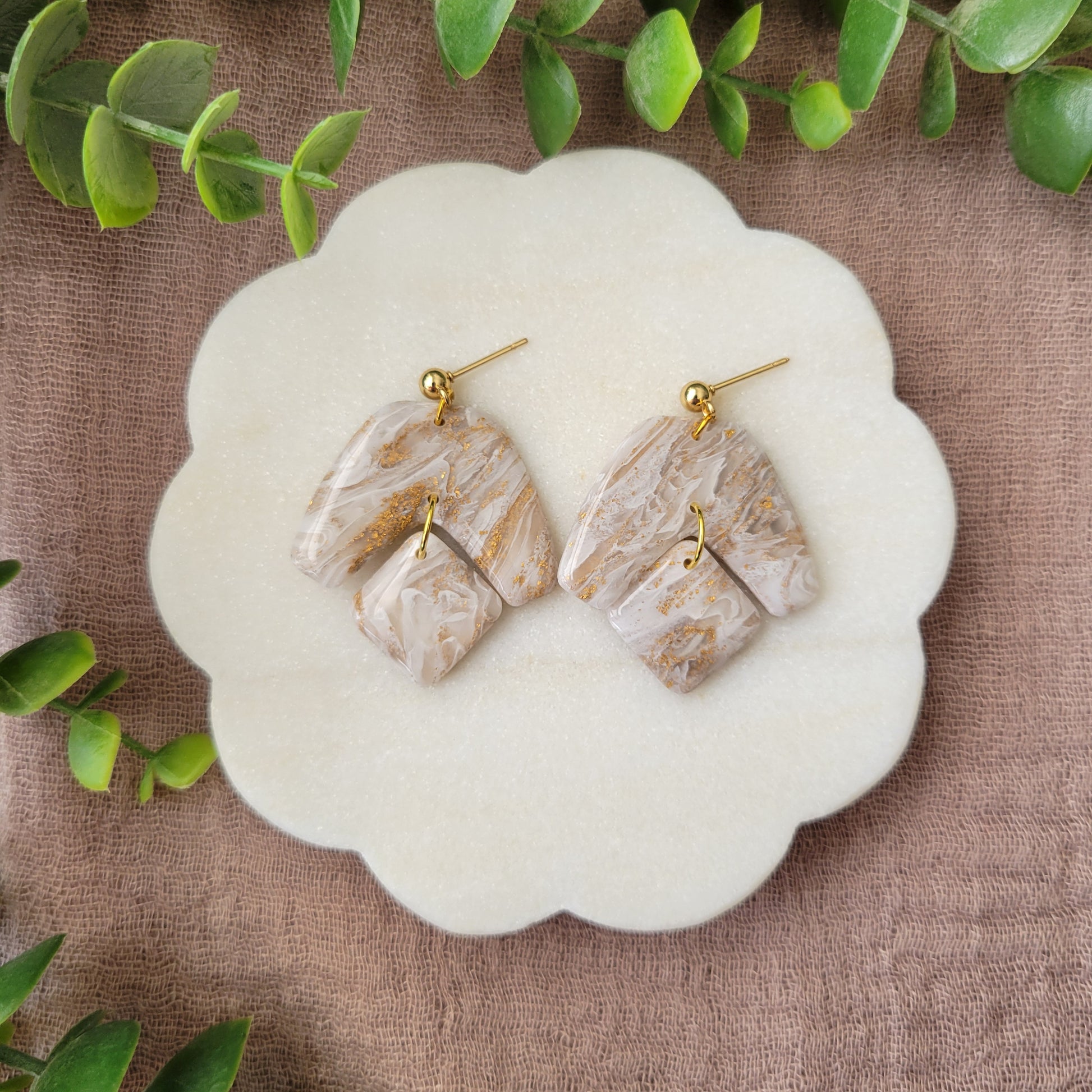Top view of a pair of white, gold marble boomerang shaped earrings displayed on a white round scalloped tray.
