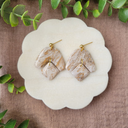 Top view of a pair of white, gold marble boomerang shaped earrings displayed on a white round scalloped tray.