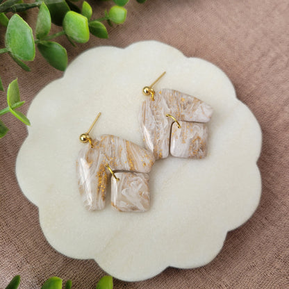 Left side view of a pair of white, gold marble boomerang shaped earrings displayed on a white round scalloped tray.