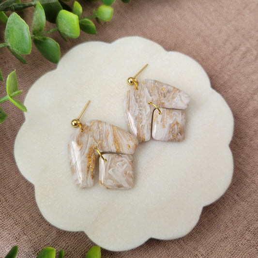 Left side view of a pair of white, gold marble boomerang shaped earrings displayed on a white round scalloped tray.