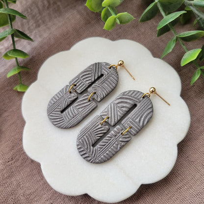 Right side view of a pair of gray earrings with art deco lines texture displayed on a white round scalloped tray.