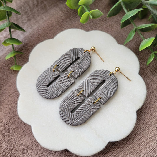 Right side view of a pair of gray earrings with art deco lines texture displayed on a white round scalloped tray.