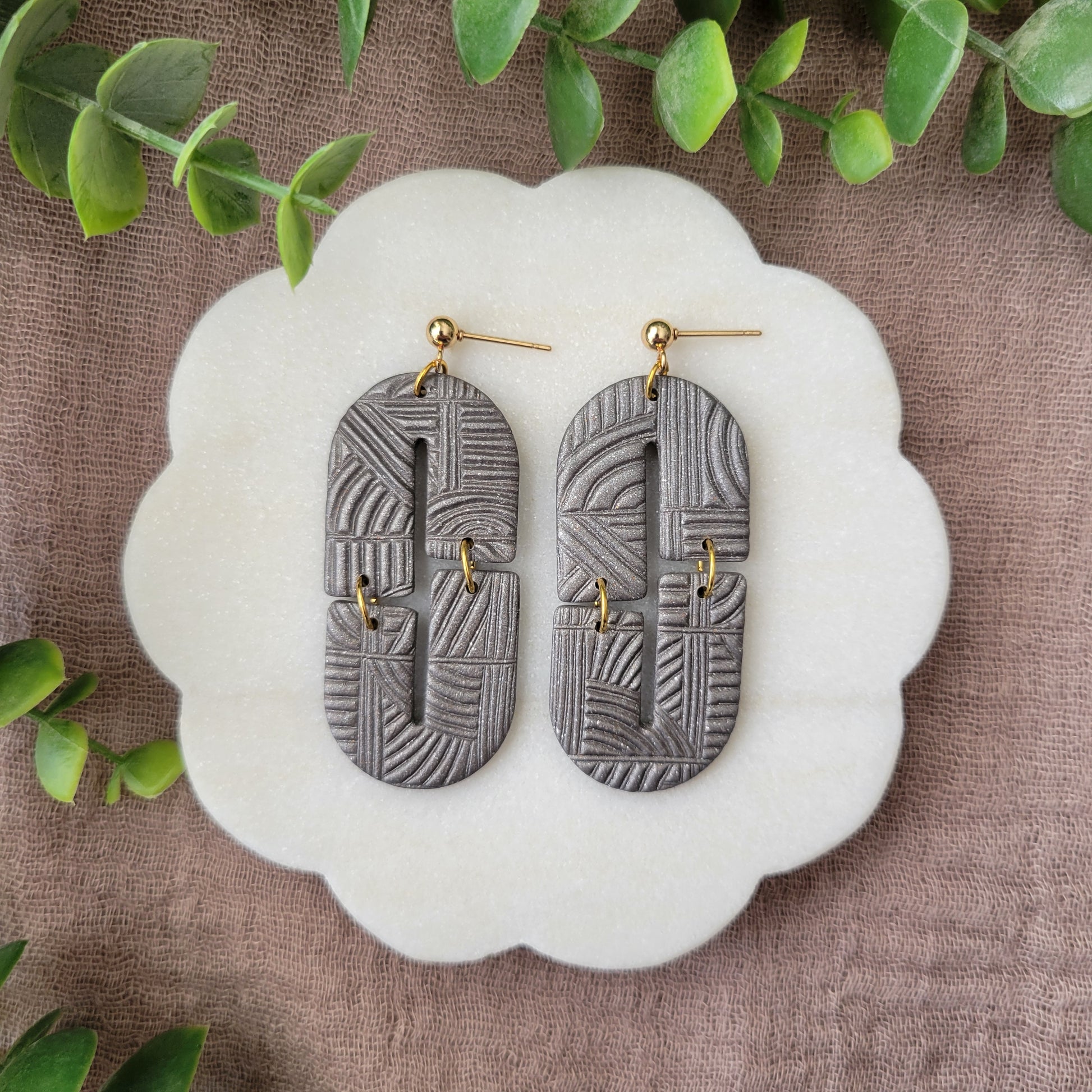 Top view of a pair of gray earrings with art deco lines texture displayed on a white round scalloped tray.