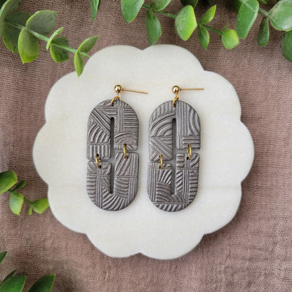 Top view of a pair of gray earrings with art deco lines texture displayed on a white round scalloped tray.
