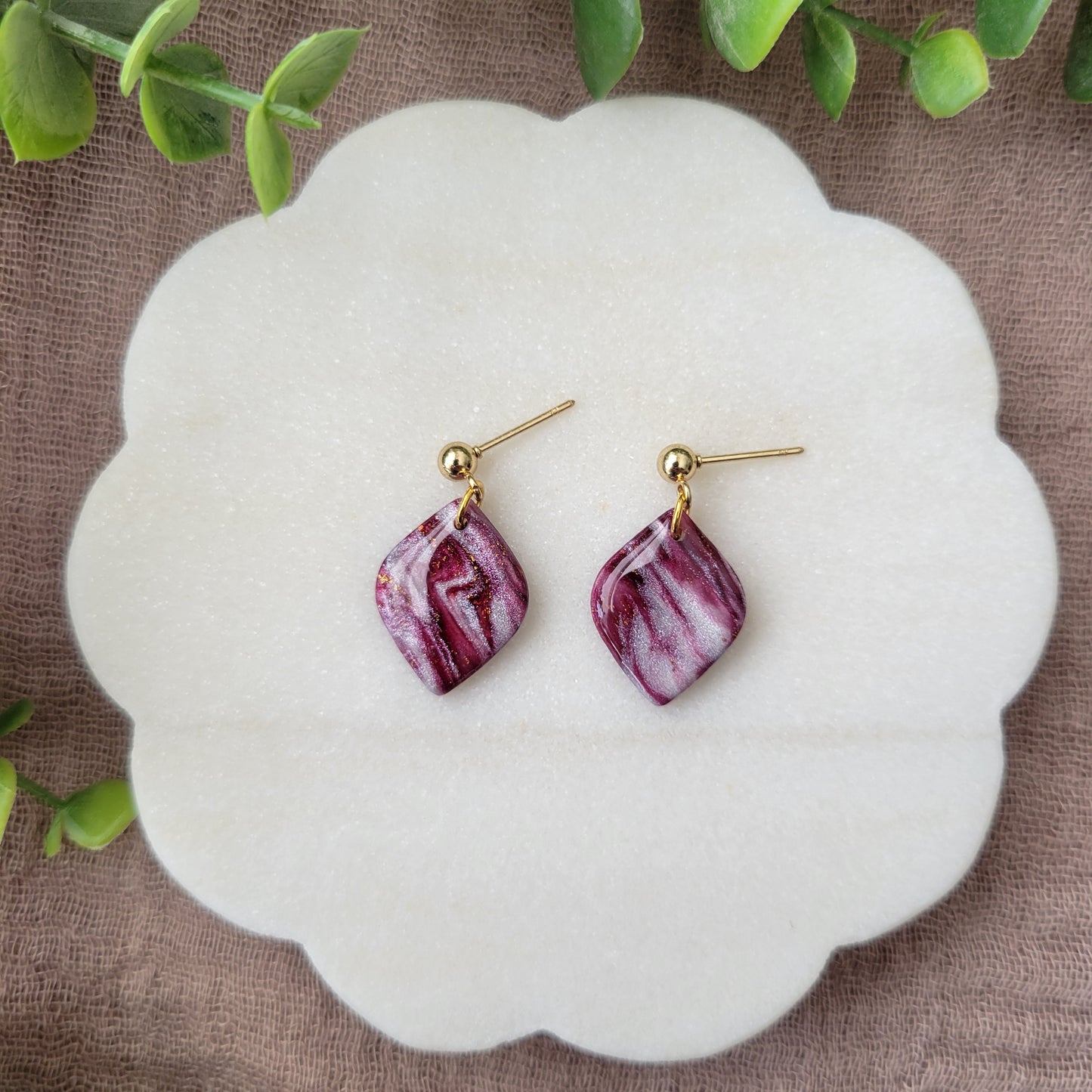 Top view of a pair of burgundy, pearl marble diamond shaped earrings displayed on a white round scalloped tray.