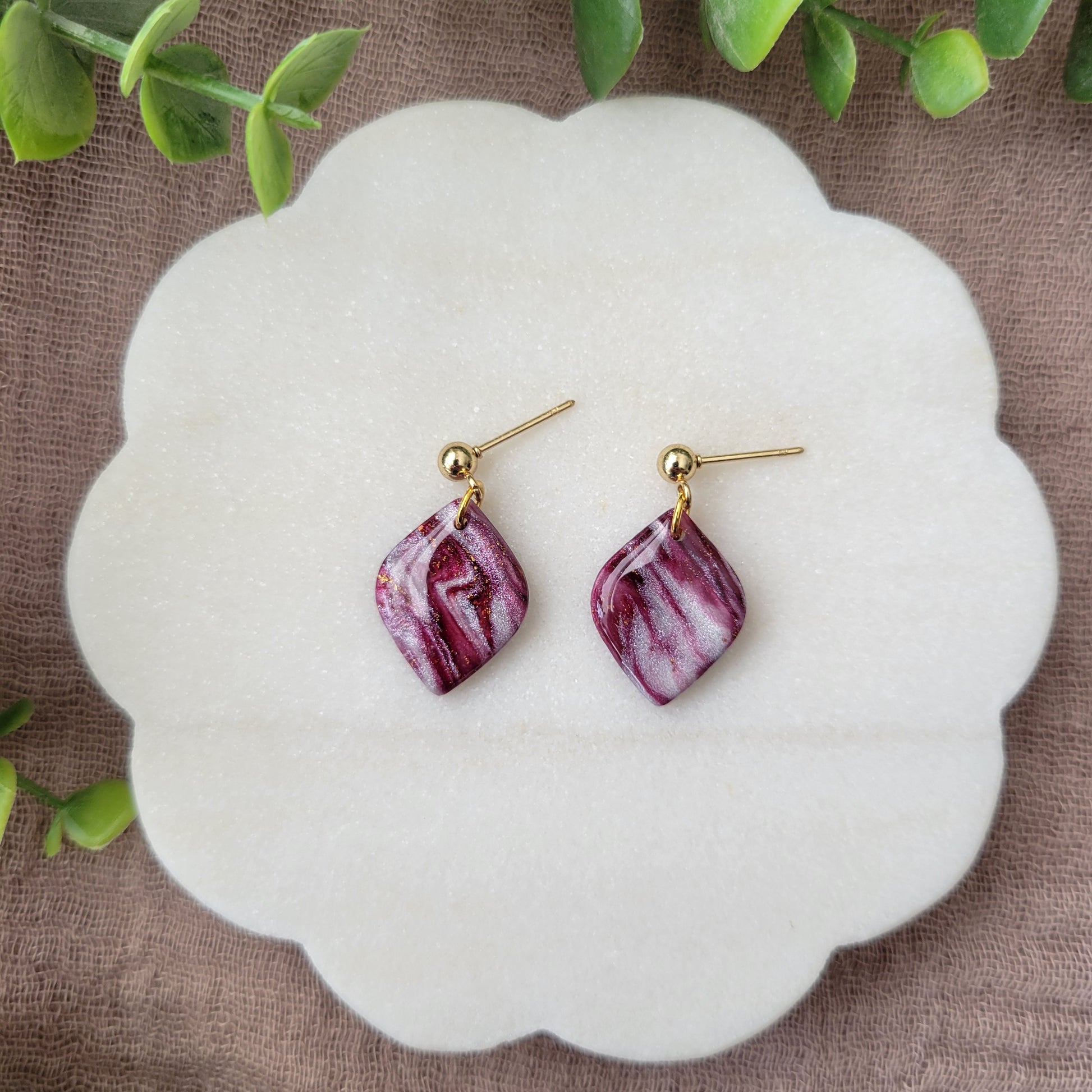 Top view of a pair of burgundy, pearl marble diamond shaped earrings displayed on a white round scalloped tray.