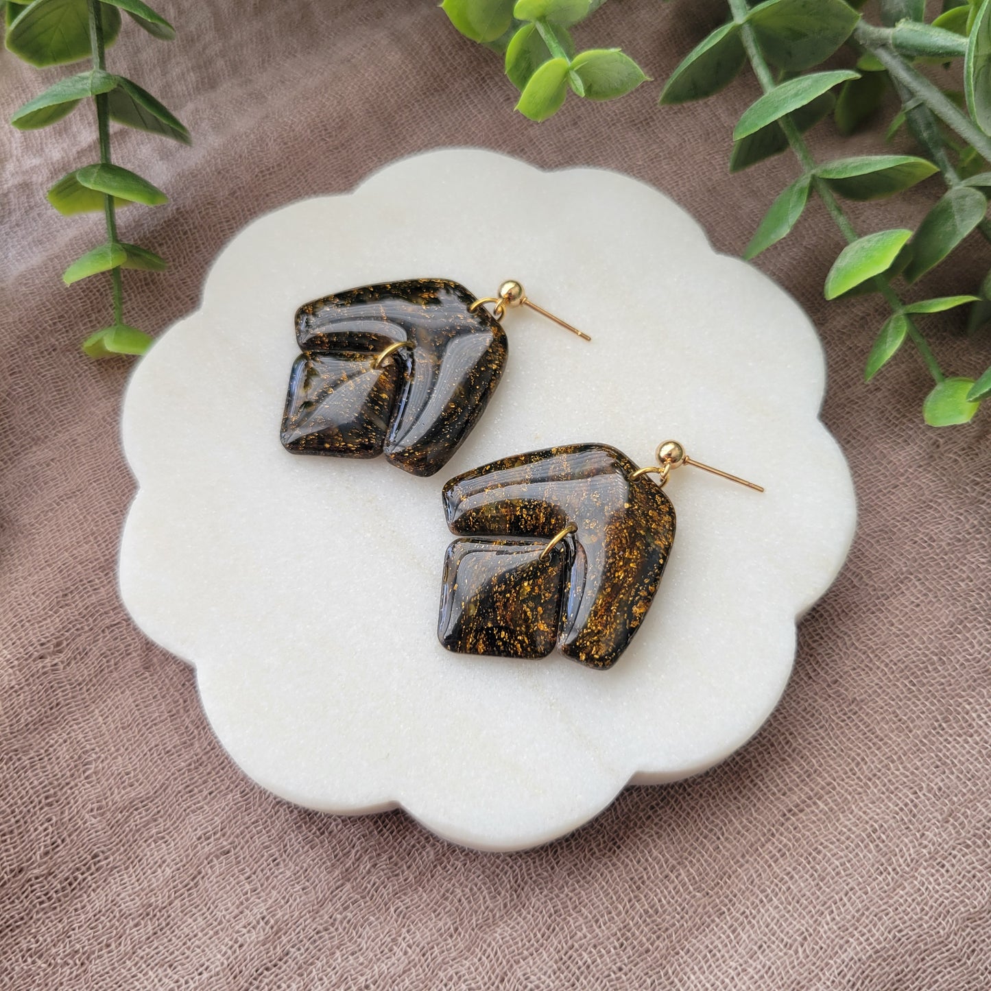 Right side view of a pair of black, gold marble boomerang shaped earrings displayed on a white round scalloped tray.
