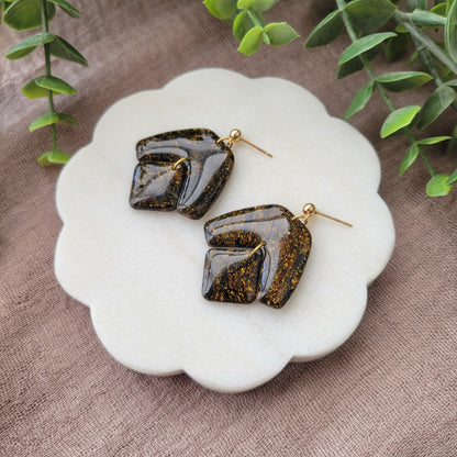 Right side view of a pair of black, gold marble boomerang shaped earrings displayed on a white round scalloped tray.