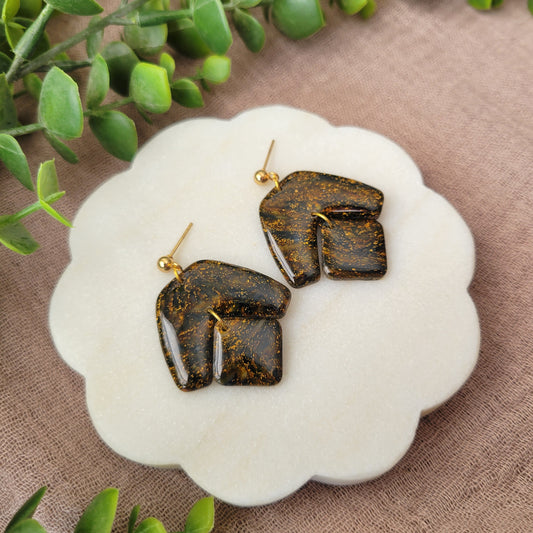 Left side view of a pair of black, gold marble boomerang shaped earrings displayed on a white round scalloped tray.