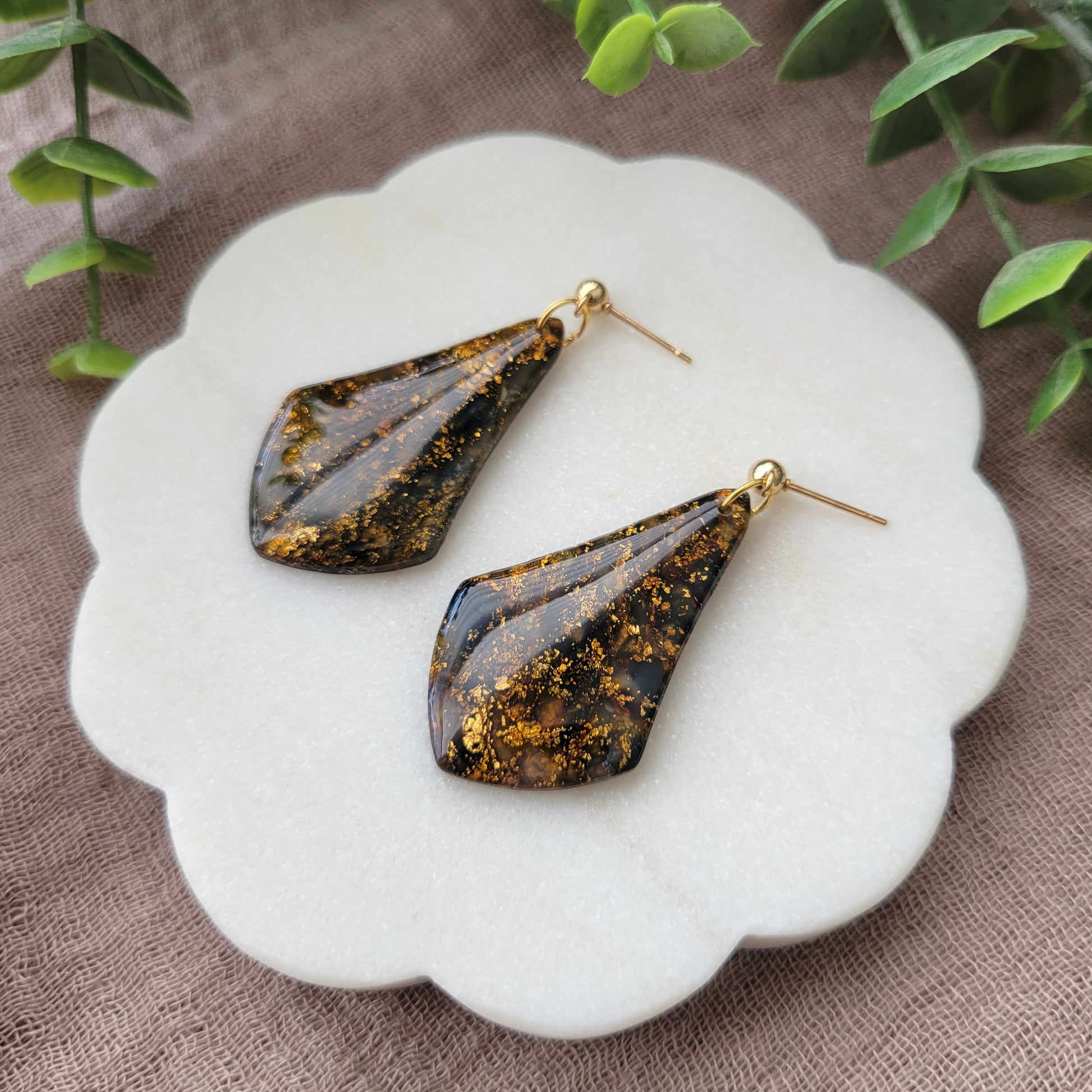 Right side view of a pair of black, gold marble wide teardrop shaped earrings displayed on a white round scalloped tray.