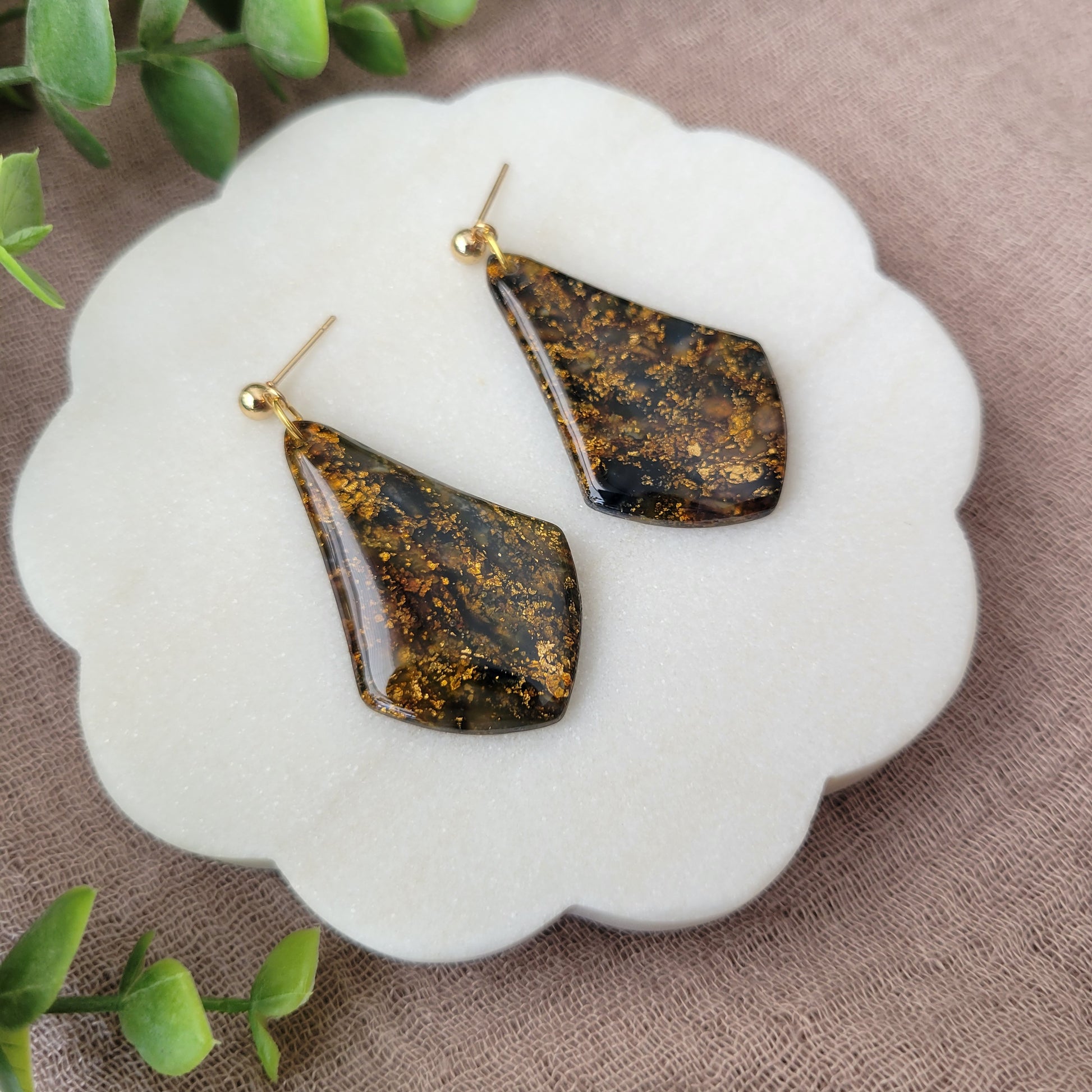 Left side view of a pair of black, gold marble wide teardrop shaped earrings displayed on a white round scalloped tray.