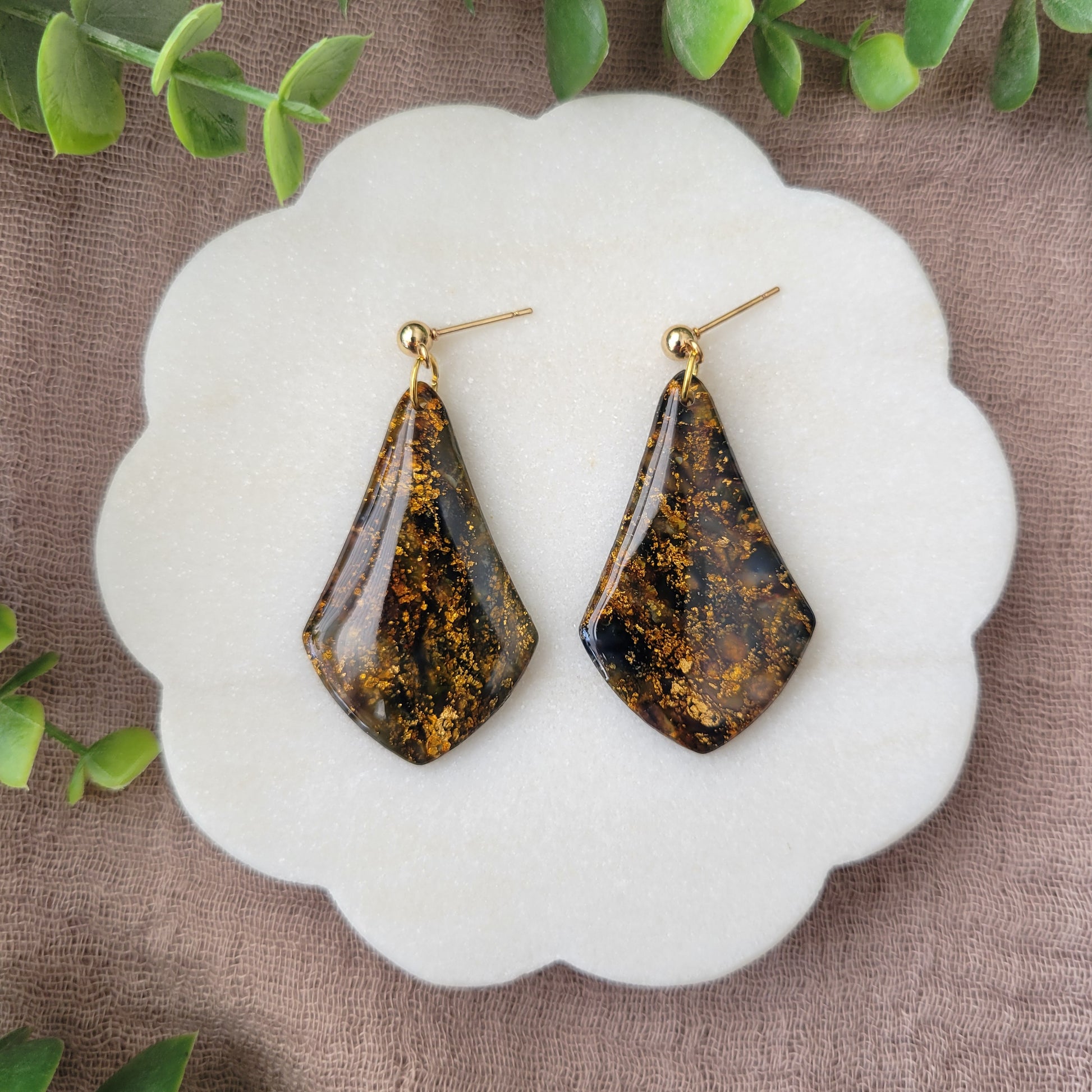 Top view of a pair of black, gold marble wide teardrop shaped earrings displayed on a white round scalloped tray.