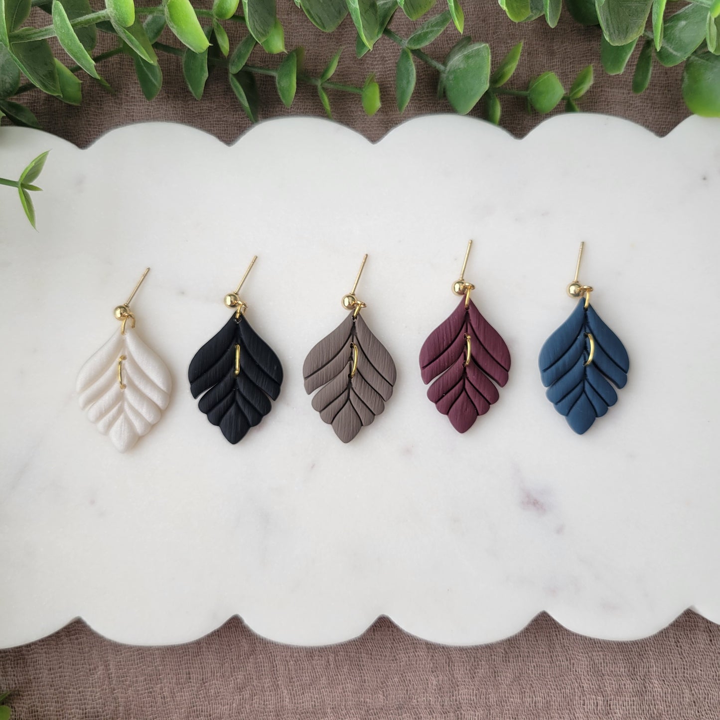 Top view of five elegant leaf shaped earrings displayed on a white scalloped tray.