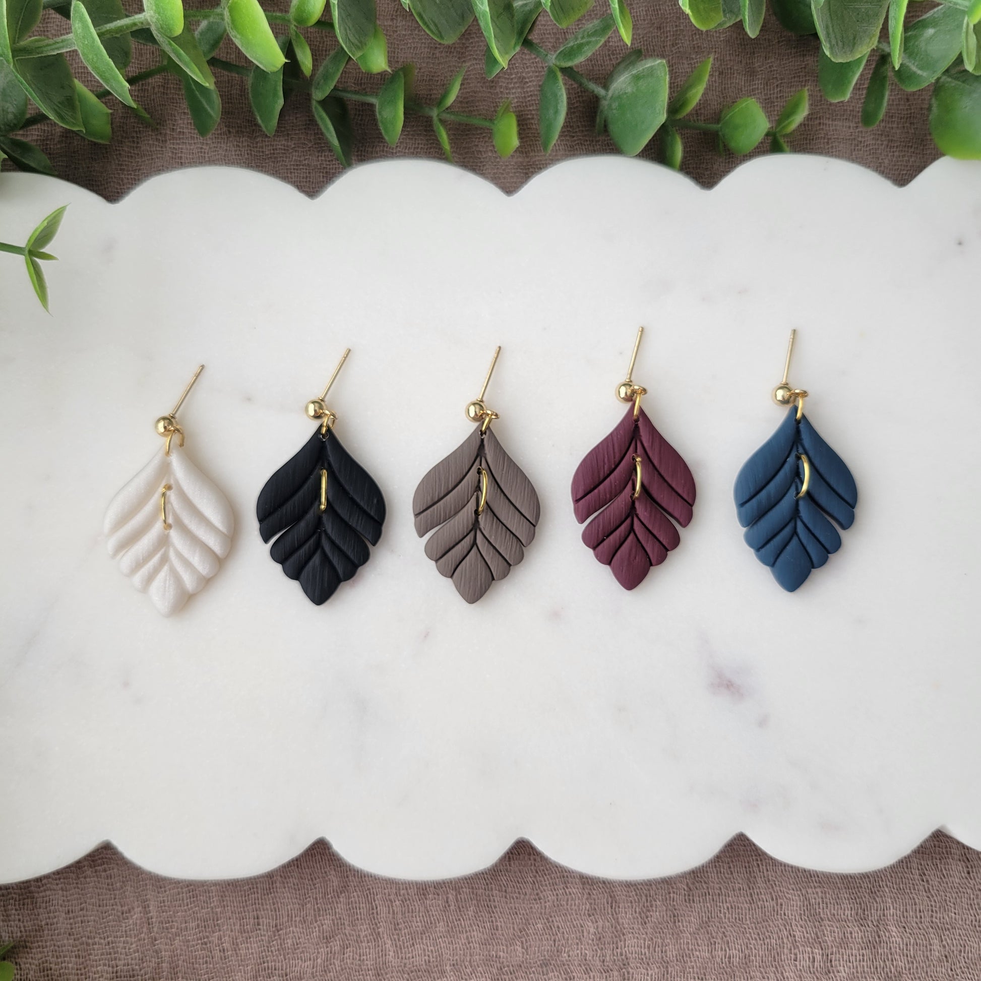 Top view of five elegant leaf shaped earrings displayed on a white scalloped tray.