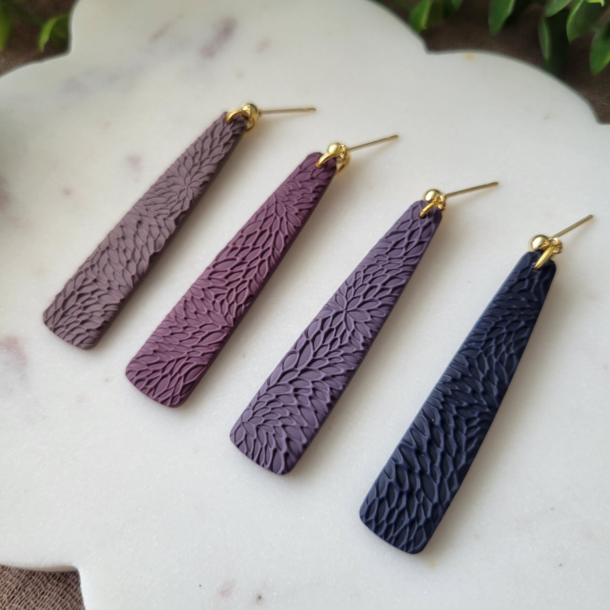 Close up of four pillar earrings with interwoven leaves texture displayed on a white scalloped tray.