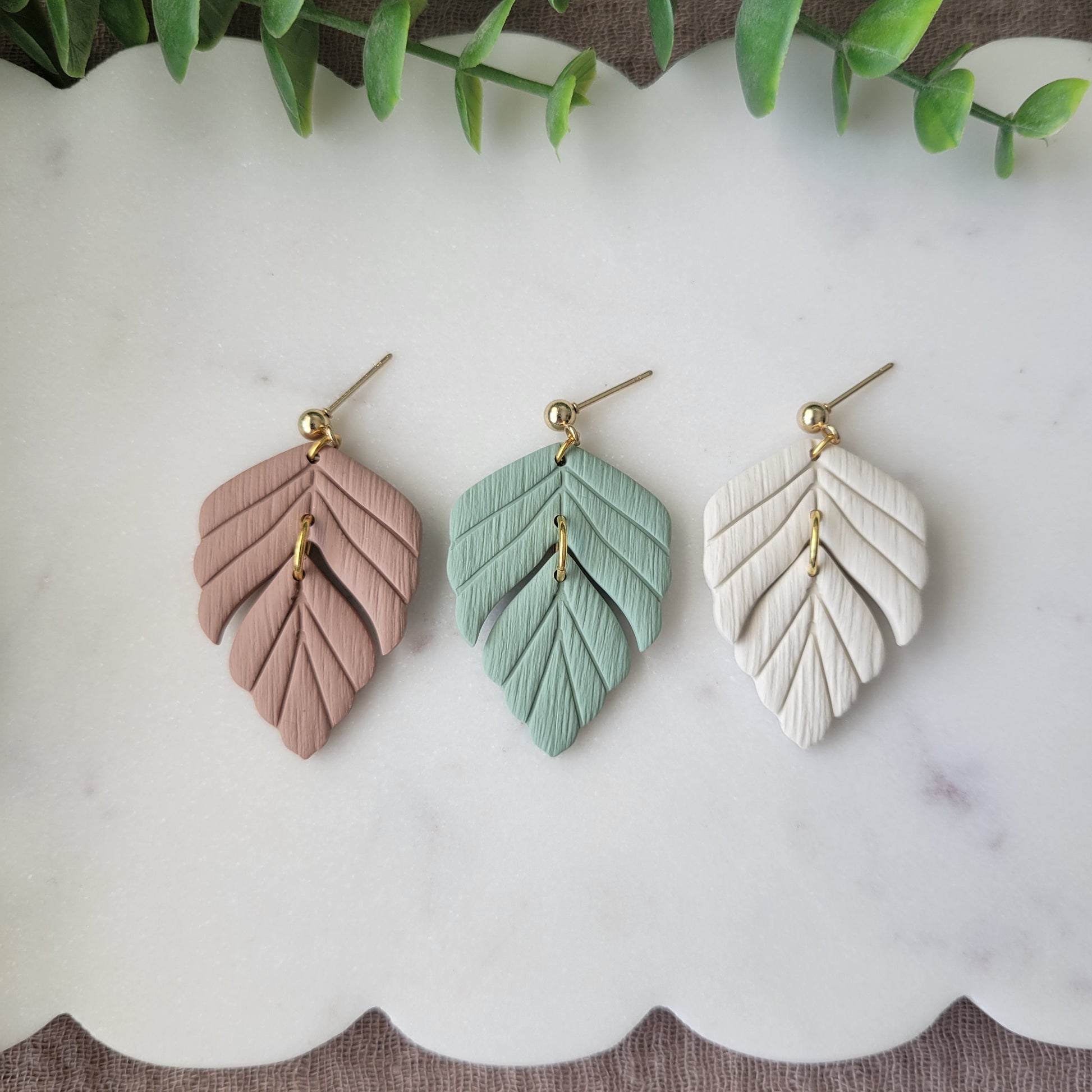 Top view of three leaf shaped earrings displayed on a white scalloped tray.