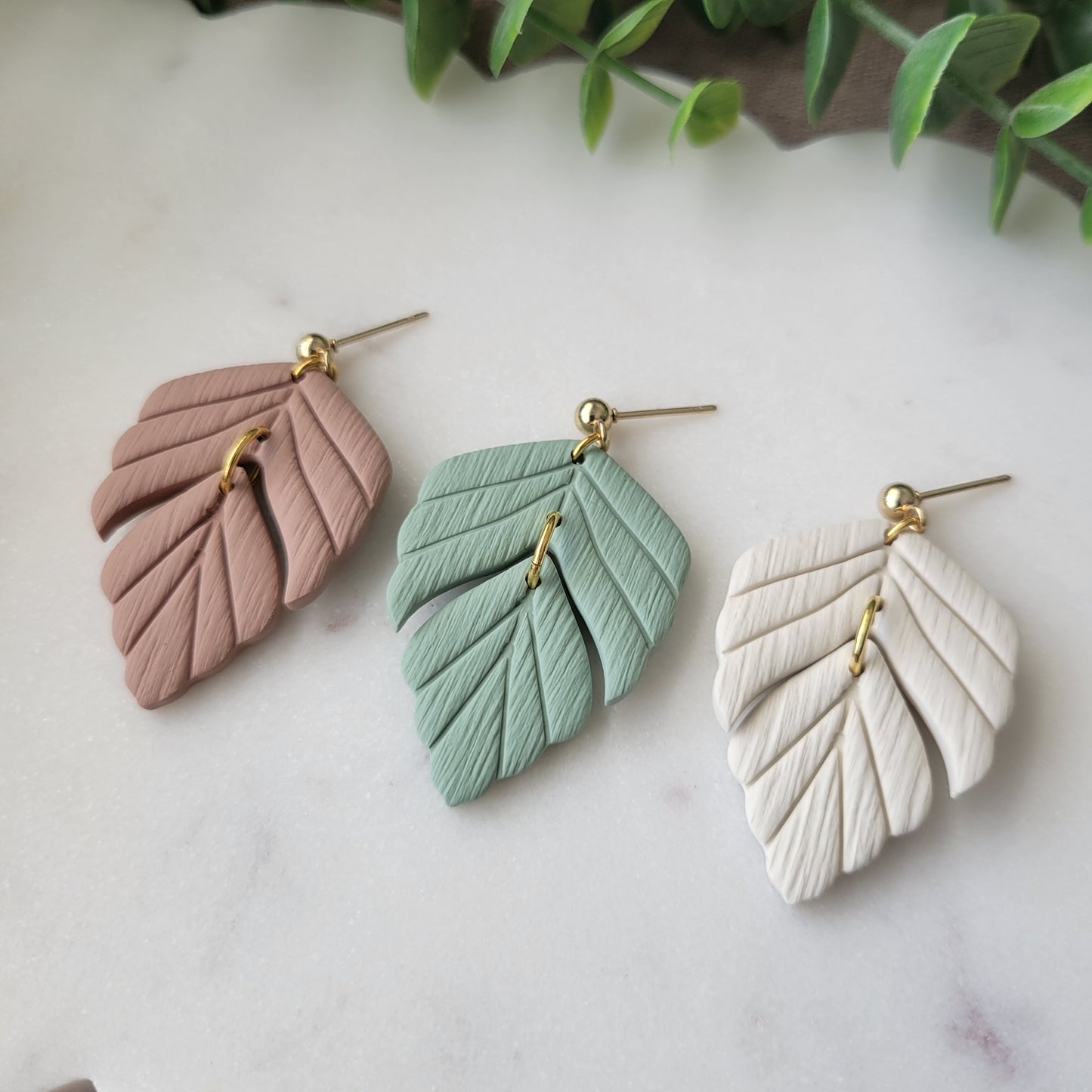Close up side view of three leaf shaped earrings displayed on a white scalloped tray.