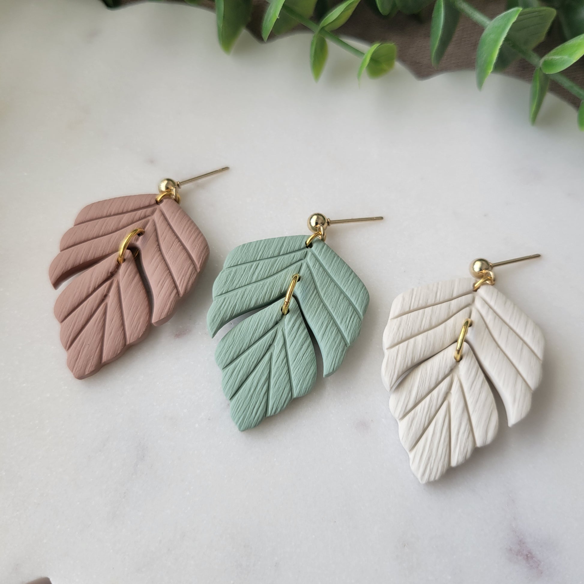 Close up side view of three leaf shaped earrings displayed on a white scalloped tray.
