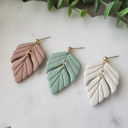 Close up side view of three leaf shaped earrings displayed on a white scalloped tray.