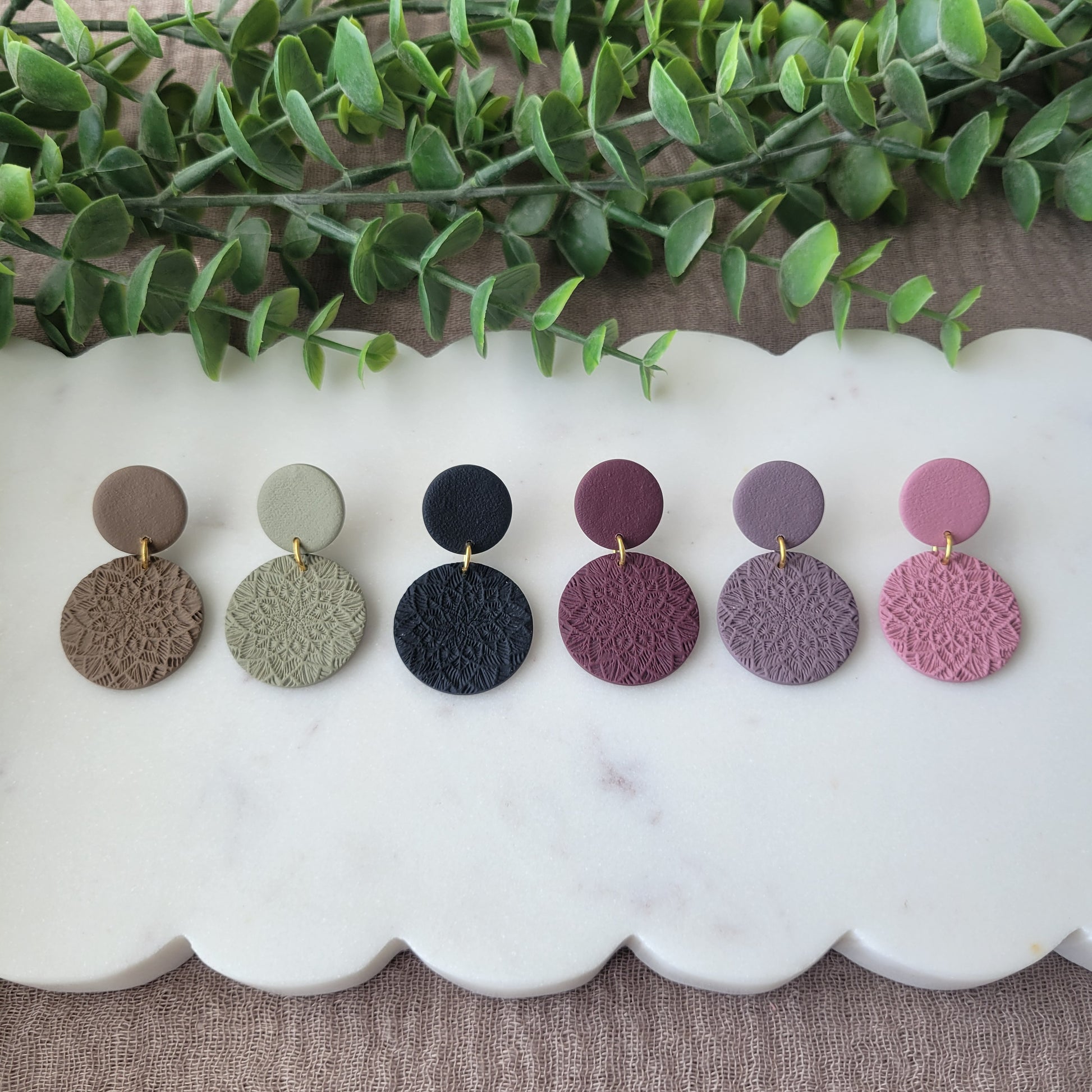 Top view of six earrings with mandala texture displayed on a white scalloped tray.