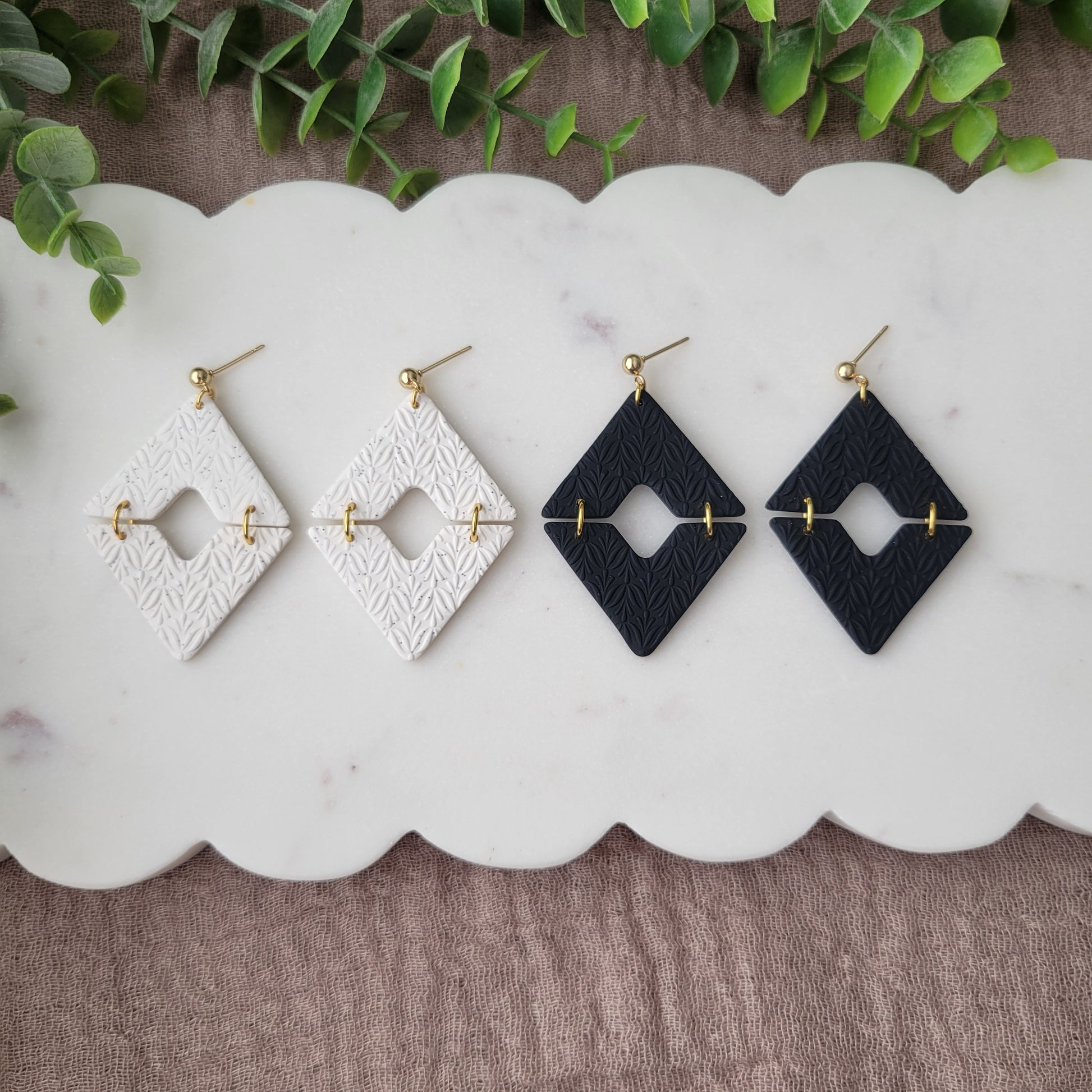 Top view of two pairs of geometric  diamond shaped earrings with art deco flower texture displayed on a white scalloped tray.