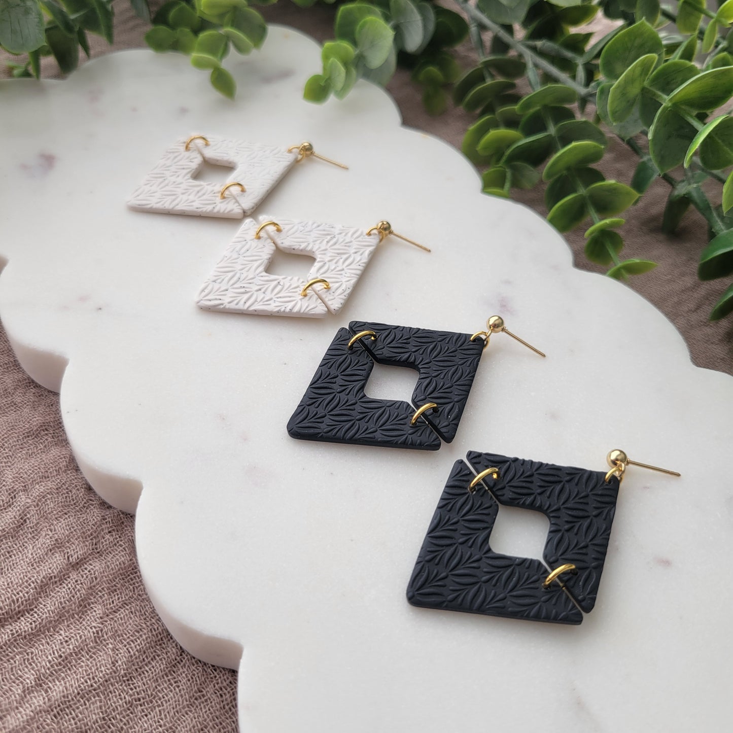 Right side view of two pairs of geometric diamond shaped earrings with art deco flower texture displayed on a white scalloped tray.