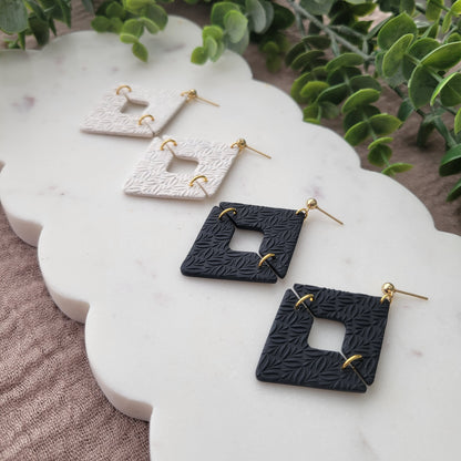 Right side view of two pairs of geometric diamond shaped earrings with art deco flower texture displayed on a white scalloped tray.