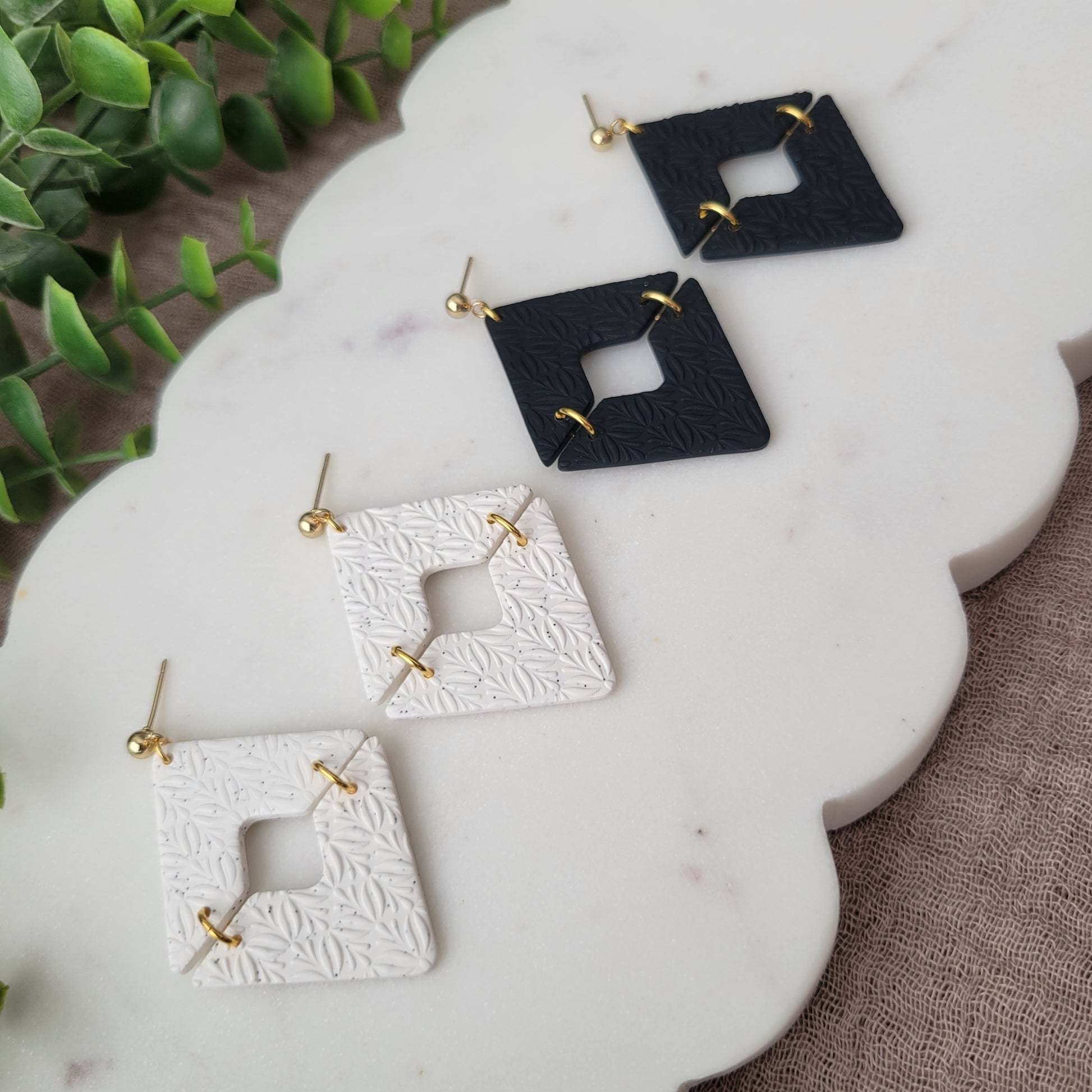 Left side view of two pairs of geometric  diamond shaped earrings with art deco flower texture displayed on a white scalloped tray.