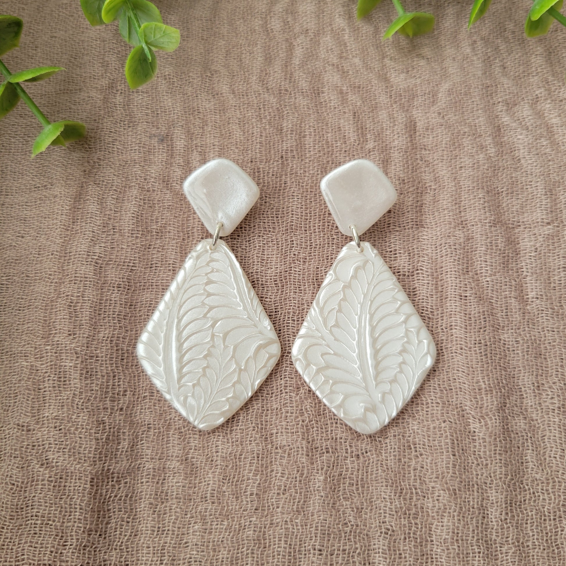 Top view of a pair of pearl geometric shaped earrings with fancy fern texture displayed on a brown cloth.