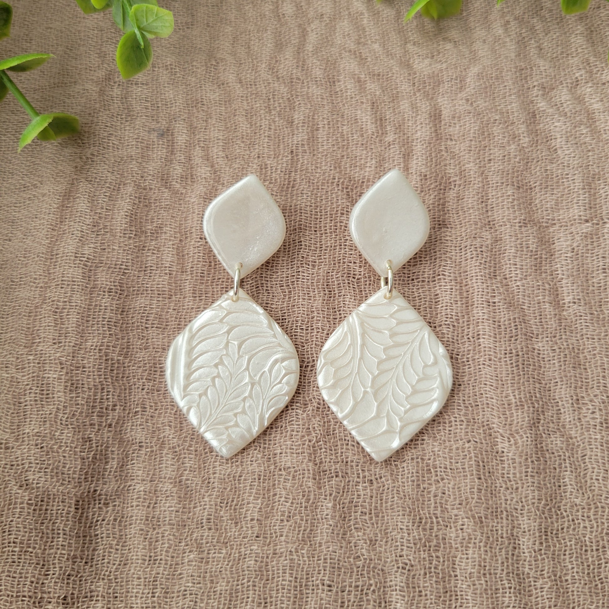 Top view of a pair of pearl diamond shaped earrings with fancy fern texture displayed on a brown cloth.