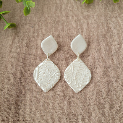 Top view of a pair of pearl diamond shaped earrings with fancy fern texture displayed on a brown cloth.