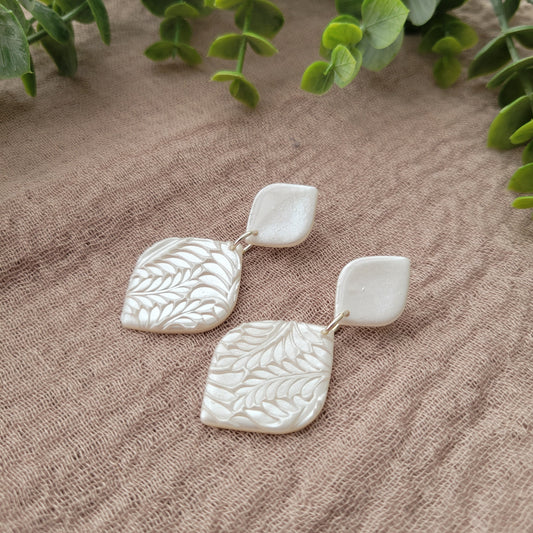 Side view of a pair of pearl diamond shaped earrings with fancy fern texture displayed on a brown cloth.