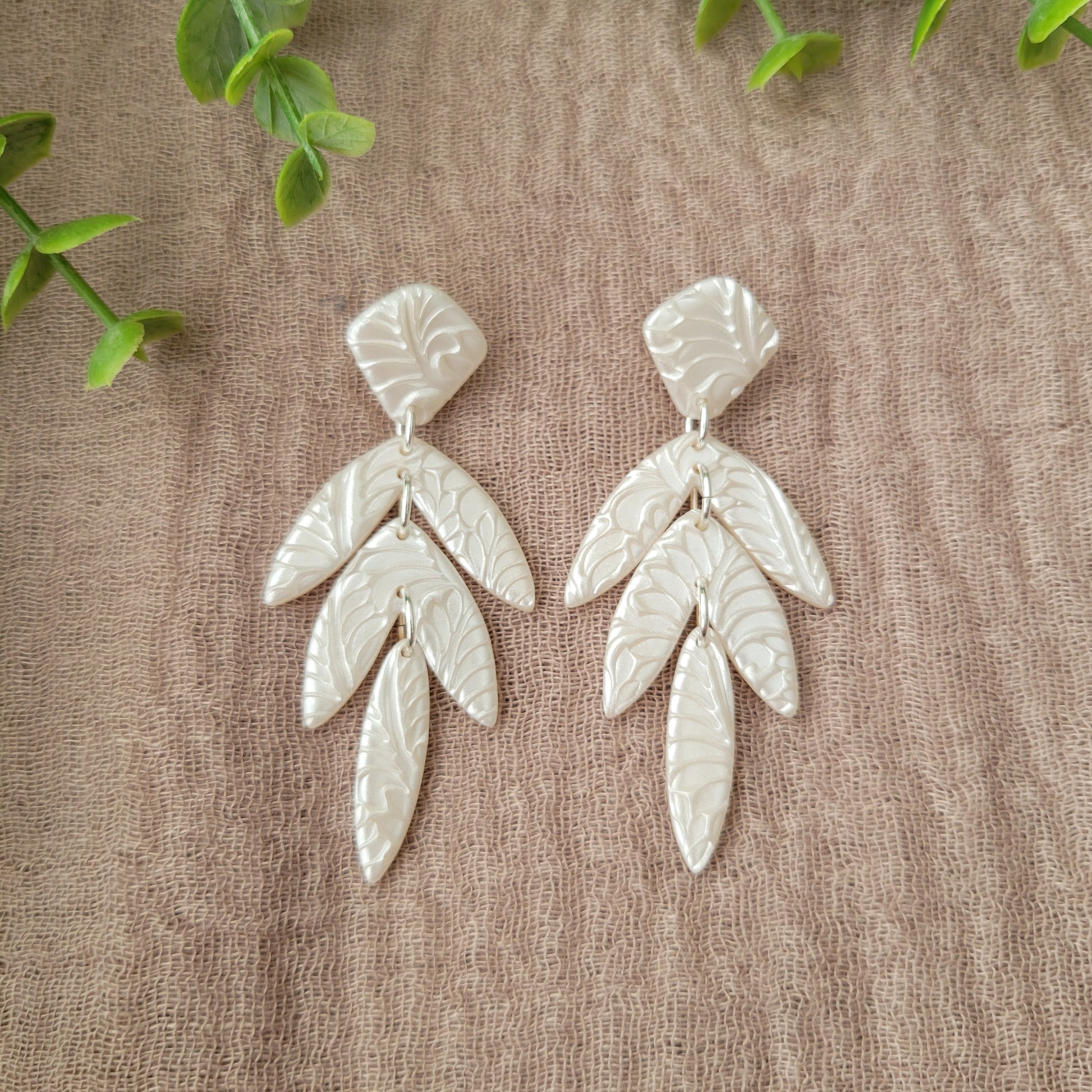 Top view of a pair of pearl long leaf shaped earrings with fancy fern texture displayed on a brown cloth.
